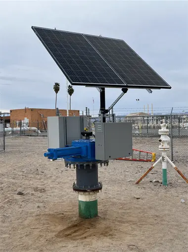 Solar panels mounted on a device in a fenced area, with industrial buildings and palm trees in the background.